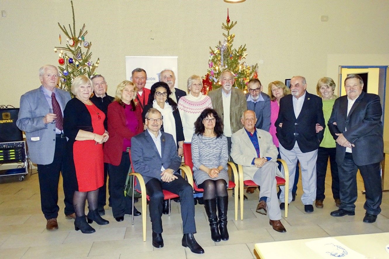 Bild: Frauen und Männer sitzen und stehen in einer Gruppe vor zwei Weihnachtsbäumen