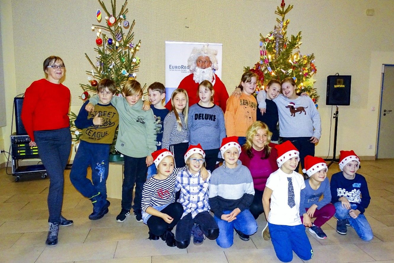 Bild: Kinder mit Nikolausmützen sitzen oder stehen in einem Raum vor zwei Weihnachtsbäumen